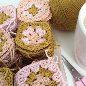 K&ouml;nnte beinhalten: Ein Stapel geh&auml;kelter Granny Squares in Rosa und Senfgelb. Die Quadrate sind zu einem Stapel gestapelt, wobei das oberste Quadrat ein rosafarbenes Zentrum und einen senfgelben Rand zeigt. Im Hintergrund befinden sich eine Kugel Senfgelb-Garn und eine rosafarbene Tasse mit Kaffee.