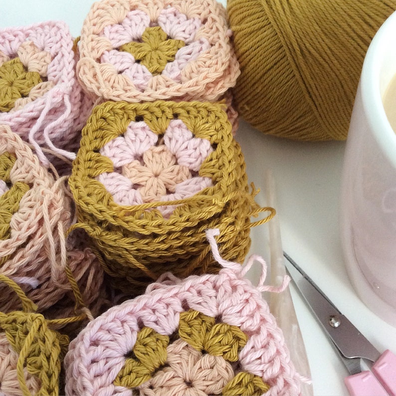 K&ouml;nnte beinhalten: Ein Stapel geh&auml;kelter Granny Squares in Rosa und Senfgelb. Die Quadrate sind zu einem Stapel gestapelt, wobei das oberste Quadrat ein rosafarbenes Zentrum und einen senfgelben Rand zeigt. Im Hintergrund befinden sich eine Kugel Senfgelb-Garn und eine rosafarbene Tasse mit Kaffee.