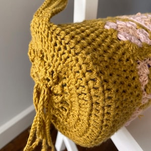 K&ouml;nnte beinhalten: Eine geh&auml;kelte Tasche in Senfgelb mit einem rosa und violetten Design. Die Tasche hat einen langen Riemen und einen Kordelverschluss.