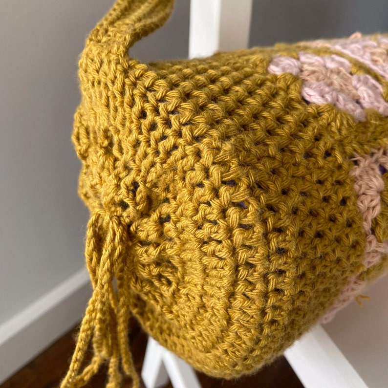 K&ouml;nnte beinhalten: Eine geh&auml;kelte Tasche in Senfgelb mit einem rosa und violetten Design. Die Tasche hat einen langen Riemen und einen Kordelverschluss.