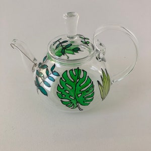 May include: A clear glass teapot with a painted design of green leaves and a monstera leaf. The teapot has a lid and a handle.
