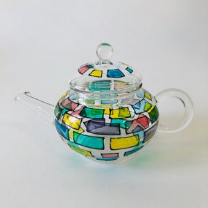 May include: A clear glass teapot with a lid and handle. The body of the teapot is decorated with colourful, rectangular shapes in shades of blue, green, yellow, pink, and purple. The teapot is designed for brewing tea.