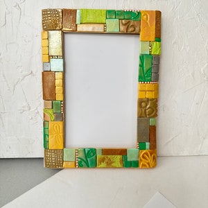 Puede incluir: Un marco de fotos con un diseño de mosaico en tonos verdes, dorados y marrones. El marco está hecho de pequeñas baldosas cuadradas que están dispuestas en un patrón aleatorio. El marco está vacío y listo para contener una foto.
