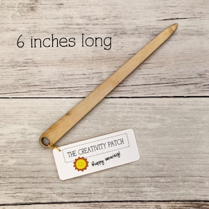 May include: A 6-inch long wooden weaving needle with a loop at one end. The needle is on a wooden surface with a white tag that says "THE CREATIVITY PATCH" and "Happy weaving!" with a yellow sun graphic.