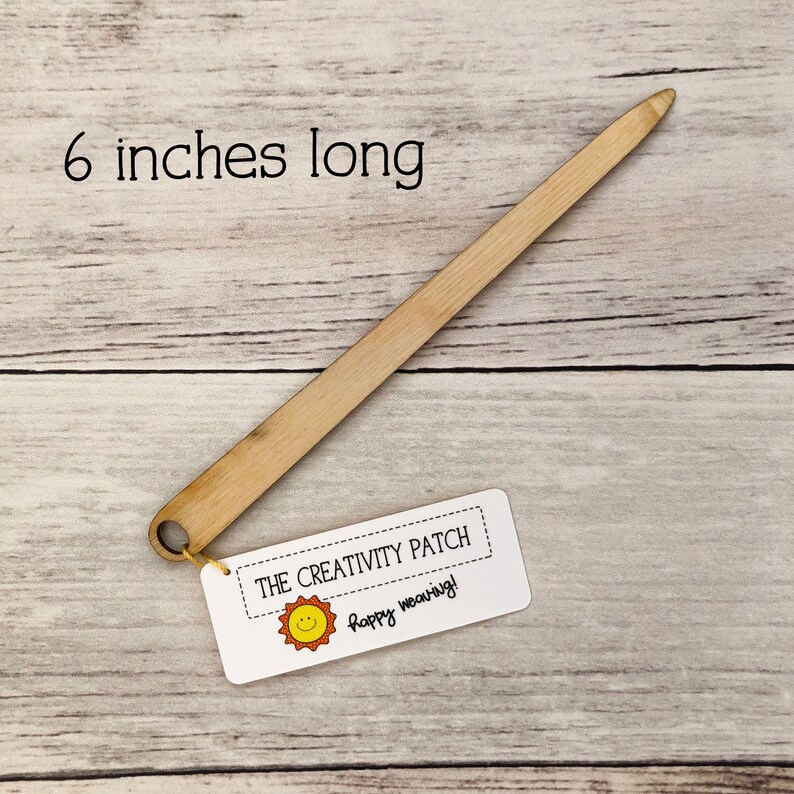 May include: A 6-inch long wooden weaving needle with a loop at one end. The needle is on a wooden surface with a white tag that says "THE CREATIVITY PATCH" and "Happy weaving!" with a yellow sun graphic.