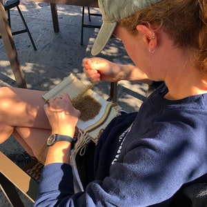 May include: A person sitting on a chair and weaving a brown, white, and yellow tapestry on a wooden loom. The person is wearing a blue shirt and a green baseball cap.