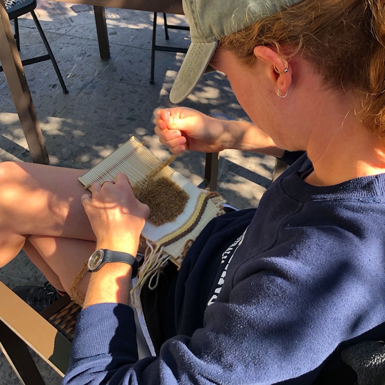 May include: A person sitting on a chair and weaving a brown, white, and yellow tapestry on a wooden loom. The person is wearing a blue shirt and a green baseball cap.