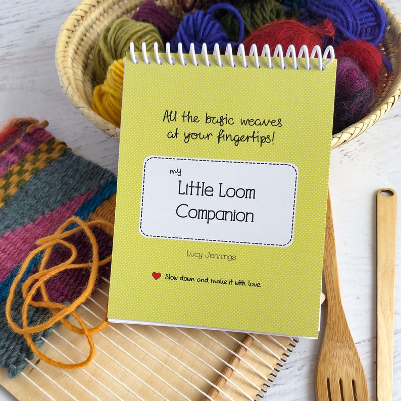 May include: A yellow booklet with a white border and polka dots. The title is "my Little Loom Companion" with the text "All the basic weaves at your fingertips!" and "Slow down and make it with love." The booklet is on a white surface with a wooden loom and yarn.