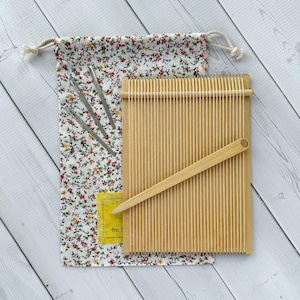 May include: A rectangular wooden weaving loom with vertical white warp threads and a wooden shuttle, accompanied by three metal needles and a drawstring fabric pouch featuring a small red and green floral pattern on a white background.