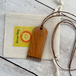 May include: A wooden weaving tool with a comb-like edge and a smiling sun design. The tool is hanging from a brown cord and is in a small, white drawstring bag. The bag has a yellow label that says "Happy weaving!"