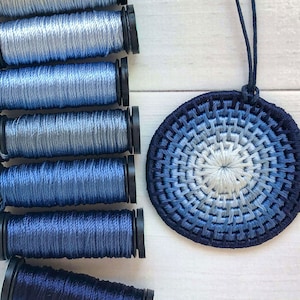 May include: A blue and white ombre circular woven pendant necklace with a black cord. The pendant is made of multiple strands of thread woven together in a circular pattern.