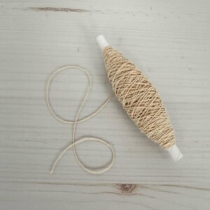 May include: A spool of natural-colored twine with a white paper core. The twine is tightly wound around the core, with a length of twine unwound and lying on the surface. The twine is a light beige color, and the background is a light wood grain.