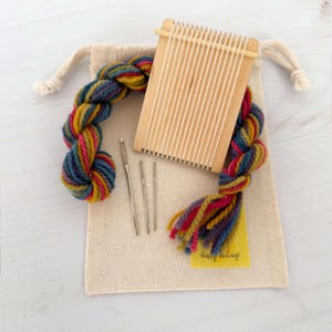 May include: A craft kit featuring a small wooden weaving loom, colorful yarn, and needles, all inside a beige drawstring bag. The yarn is a mix of blue, yellow, red, and pink. A small yellow tag with the text "Happy weaving!" is included.