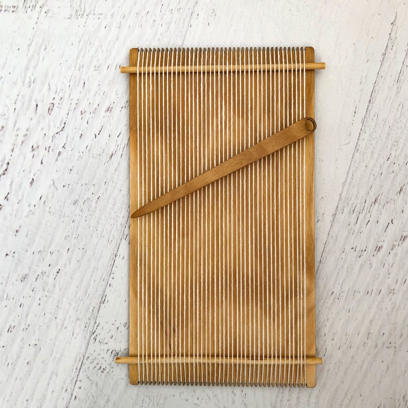 May include: A wooden loom with a wooden shuttle, used for weaving yarn or string. The loom is made of natural wood and has a white string threaded through it.