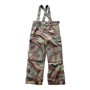 May include: Camouflage pattern overalls with suspenders. The pants feature a mix of green, brown, and tan colors. The overalls have two front pockets and button closures at the ankles. The suspenders are attached to the waistband.