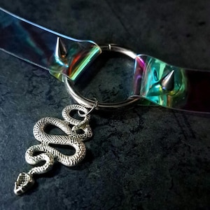 May include: A choker necklace with a silver snake pendant. The choker is made of iridescent, clear plastic with silver metal accents and a ring clasp. The necklace has two silver spikes on the ends of the plastic band.