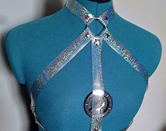 May include: A clear, holographic harness with silver-tone hardware. The harness features a choker-style collar, straps, and a circular design with a black center. Small star-shaped accents are placed on the harness.