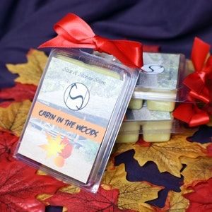 May include: A set of wax melts in clear plastic containers with a red ribbon. The wax melts are labeled "Cabin in the Woods" and "Soar at Siren Stone".