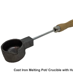 Könnte beinhalten: Ein Schmelztiegel aus Gusseisen mit Holzgriff. Der Topf ist dunkelgrau und hat eine abgerundete Schalenform. Der Griff ist mit einer Metallstange befestigt und besteht aus hellfarbenem Holz. Der Text "BLACK BARN DESIGN" befindet sich links.