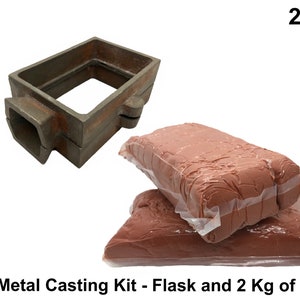 May include: A metal casting kit featuring a rectangular flask and two packages of sand, each weighing 2 Kg. The flask is a dark, weathered metal. The sand is a reddish-brown color, packaged in clear plastic. The text "Metal Casting Kit - Flask and 2 Kg of Sand" is visible.