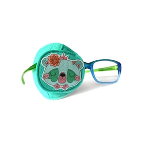 May include: A turquoise eye patch with a cartoon bear design and a green elastic strap. The eye patch is paired with blue-framed glasses.