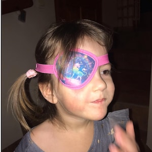 Può includere: Una bambina che indossa una benda per l'occhio rosa con un disegno di unicorno. La benda per l'occhio è fissata con un elastico rosa.