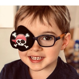 May include: A young boy wearing blue glasses and a black eye patch with a pink skull and crossbones design.