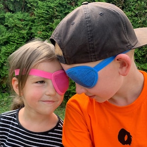 Può includere: Due bambini con una benda sull'occhio, una rosa e una blu, si guardano. Il bambino a sinistra indossa una camicia a righe bianche e nere. Il bambino a destra indossa un berretto da baseball nero e una maglietta arancione con un disegno nero.