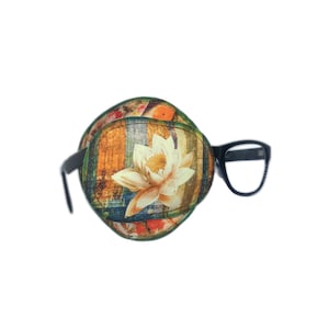 Könnte beinhalten: Ein schwarzes Augenpflaster mit einem Blumendruck, der eine weiße Blume mit orangefarbenen und gelben Akzenten zeigt. Das Augenpflaster ist an einer schwarzen Brille befestigt.