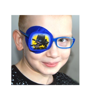 Pode incluir: Um tapa-olho azul com uma imagem de desenho animado do Batman. O tapa-olho está sendo usado por uma criança que usa óculos de armação azul.