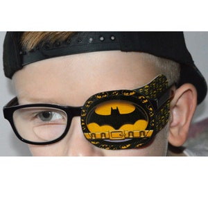 May include: A black and yellow eye patch with a Batman logo. The eye patch is worn by a child wearing glasses and a black baseball cap.