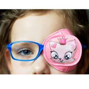 Pode incluir: Uma menina pequena usando óculos azuis com um tapa-olho rosa com um gato de desenho animado com uma coroa. O tapa-olho é feito de tecido macio e tem um design bonito.