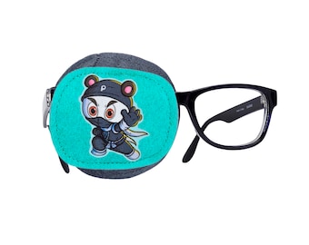 May include: A black eye patch with a cartoon ninja bear design on a turquoise background. The eye patch is attached to a pair of black framed glasses.