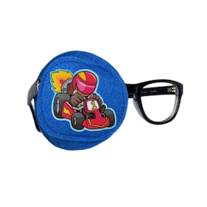 Puede incluir: Parche para el ojo azul con una imagen de dibujos animados de un piloto de carreras con un casco rojo conduciendo un coche de carreras rojo con una llama amarilla. El parche para el ojo está unido a unas gafas con montura negra.