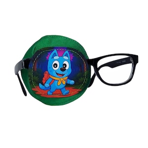 Könnte beinhalten: Eine grüne Augenklappe mit einer Cartoon-Hunde-Illustration, befestigt an einer Brille mit schwarzem Rahmen. Der Hund ist blau mit einem roten Rucksack und einem gelben Schal. Die Brille hat eine rechteckige Form.