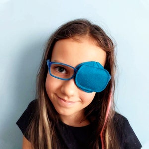 May include: A teal eye patch is worn over the left eye, attached to blue eyeglasses. The eye patch is made of soft material and is stitched around the edges. The background is a solid light blue.