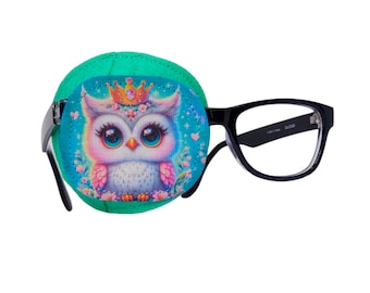 May include: A green eye patch with a colorful cartoon owl wearing a crown. The eye patch is attached to a pair of black framed glasses.