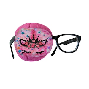 Op de afbeelding: Roze ooglap met een eenhoornontwerp met een kroon, bloemen en sterren. De ooglap is bevestigd aan een zwarte bril.