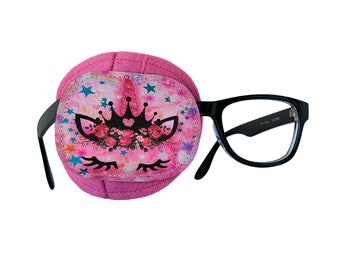 May include: Pink eye patch with a unicorn design featuring a crown, flowers, and stars. The eye patch is attached to black framed glasses.
