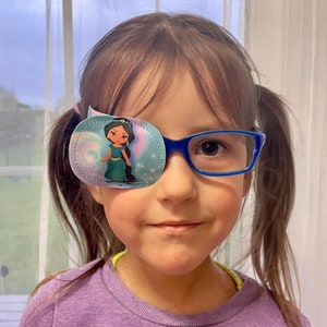 Può includere: Una bambina con gli occhiali e una benda per l'occhio blu con un personaggio di una principessa. La benda per l'occhio è decorata con uno sfondo blu chiaro e viola con stelle.