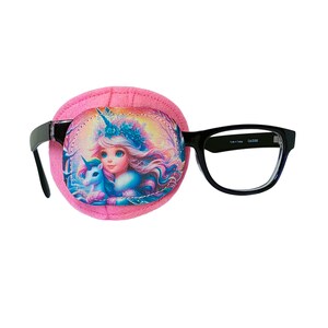Puede incluir: Parche para el ojo rosa con una ilustración de unicornio y hada. El parche está unido a unas gafas con montura negra.