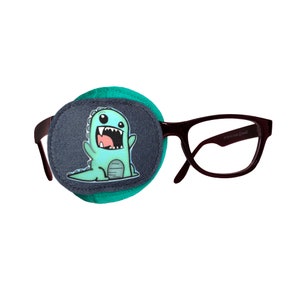 May include: A teal blue eye patch with a cartoon dinosaur graphic. The eye patch is attached to a pair of black framed glasses.