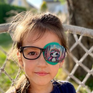 Puede incluir: Una niña pequeña con gafas de montura negra y un parche de ojo verde con un diseño de unicornio y bosque.