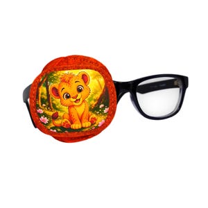 Puede incluir: Un parche ocular circular de tela con una colorida ilustración de un cachorro de león de dibujos animados sentado en un bosque con flores rosas. El parche está sujeto a unas gafas de montura negra con lentes transparentes para la visión.