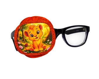 May include: A circular fabric eye patch featuring a colorful illustration of a cartoon lion cub sitting in a forest with pink flowers. The patch is attached to a pair of black-framed eyeglasses with clear lenses, designed for vision correction.