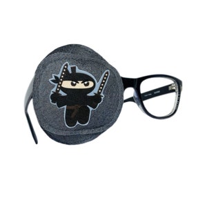 May include: A gray eye patch with a cartoon ninja design. The eye patch is attached to a pair of black framed glasses.