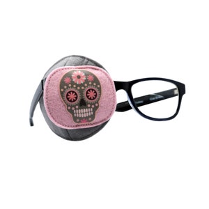 May include: A gray eye patch with a pink felt skull design, decorated with flowers, is attached to a pair of black eyeglasses. The eye patch is designed to cover one eye. The glasses have a black frame.