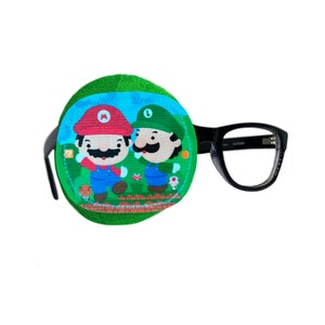 Può includere: Un cerotto per l'occhio verde con un'immagine di cartone animato di Mario e Luigi su uno sfondo verde. Il cerotto per l'occhio è attaccato a un paio di occhiali con montatura nera.