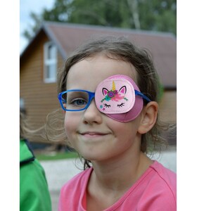 May include: A young girl wearing blue glasses and a pink eye patch with a unicorn design. The eye patch has a pink background with a white unicorn with a gold horn, pink eyelashes, and a floral crown.