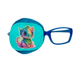 Op de afbeelding: Een blauwe ooglap met een kleurrijk cartoon-draakontwerp. De lap is bevestigd aan een paar blauwe plastic brillen.